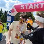 Na zdjęciu widać uczestników biegu na starcie, pod różowo-białym łukiem. Osoba w jasnej koszulce otrzymuje medal. W tle są drzewa i ludzie obserwujący wydarzenie.