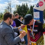 Mężczyzna w marynarce wręcza medale biegaczowi w czapce i kolorowej koszulce. W tle zielone drzewa i inne osoby. Nad nimi widoczny jest kolorowy balon z napisem.