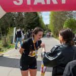 Zawodniczka w czarnej koszulce otrzymuje medal pod różowym łukiem startowym. W tle zieleń i kilku biegaczy. Ciepły, słoneczny dzień.