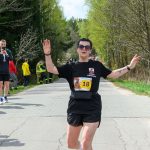 Biegaczka przekracza linię mety na leśnej drodze. Ubrana na czarno, w tle zielone drzewa. Na koszulce numer 18.