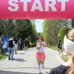 Zawodniczka w różowej spódnicy biegnie przez metę pod różowym łukiem z napisem START. W tle jest las. Obserwatorzy stoją przy drodze.