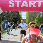 Zdjęcie przedstawia biegacza w białej koszulce pod różowym napisem START. Tło to zielone drzewa i niebo. Obok są inni uczestnicy i kibice.