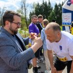 Na zdjęciu mężczyzna w okularach wręcza medal biegaczowi w białej koszulce na tle zieleni. W tle kilka osób i dmuchany łuk. Kolory są jasne, przeważają niebieski, biały i zielony.