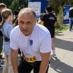 Biegacz w białej koszulce odpoczywa po biegu. W tle widać balon z napisem „Ale czas!” i ludzi. Tło jest zielone, z drzewami.