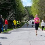 Kobieta w różowej koszulce biegnie podczas wyścigu na wiejskiej drodze. Ścieżka otoczona zielonymi drzewami i kibicami. Słońce świeci, a ona ma numer startowy 23.