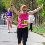 Biegaczka w różowej koszulce numer 23 biegnie z uniesionymi rękami. W tle widoczni inni zawodnicy. Scena na tle zielonych drzew.