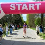 Uczestniczka biegu przekracza różowo-białą bramę startową, otoczoną drzewami. Po lewej stronie widzowie klaskają, motywując biegaczy. Na tablicy czas 0:33:36.