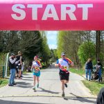 Biegacze startują spod różowego łuku startowego. Tło to zielone drzewa i jasne niebo. Naokoło stoją kibice.