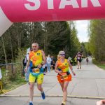 Dwoje biegaczy w kolorowych strojach przekracza linię startu pod różowym łukiem. W tle zielone drzewa i niebo. Na drodze inni uczestnicy.