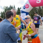 Na zdjęciu znajdują się uczestnicy biegu w żółtych strojach, stojący przed kolorowym łukiem z napisem start. W tle widoczne są drzewa i grupa osób. Atmosfera jest pełna energii i radości.