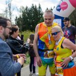 Na zdjęciu para biegaczy w kolorowych strojach z medalami. W tle widoczne są drzewa i baner startowy. Pogoda jest słoneczna, co dodaje energii wydarzeniu.