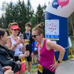 Kobieta w różowej koszulce i czarnych okularach biega maraton. W tle ludzie z numerami startowymi, nadmuchana brama i drzewa. Uczestnicy cieszą się i robią zdjęcia.