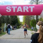 Biegacz przekracza linię startu, różowy balon z napisem START. W tle las i kibice, niebo częściowo zachmurzone.