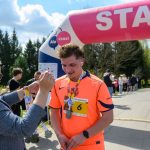 Biegacz w pomarańczowej koszulce otrzymuje medal. Tło to zielone drzewa i niebo z chmurami. Nad nim widoczny jest łuk startowy.