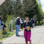 Biegaczka w czarnej koszulce i fioletowych legginsach uśmiechnięta przekracza linię mety. W tle zielone drzewa i kilku obserwatorów. Słoneczny dzień i jasno szara droga.