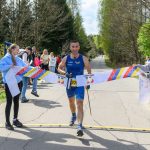 Mężczyzna w niebieskim stroju biegowym przekracza linię mety. W tle las i kibice. Jasne, słoneczne niebo.