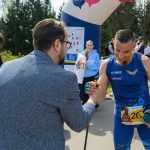 Na zdjęciu mężczyzna w niebieskim stroju sportowym na zawodach. W tle zielone drzewa i ludzie na chodniku. Scena na świeżym powietrzu.