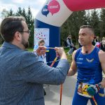 Na zdjęciu mężczyzna w niebieskim stroju biegowym otrzymuje medal. W tle znajduje się brama z napisem Ale czas! i drzewa. Obok widoczna jest grupa kibiców.