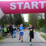 Dwie kobiety biegną pod różowym łukiem startowym na asfaltowej drodze. W tle zielone drzewa i widzowie. Kobieta w czarnej koszulce numer 1, druga w niebieskiej numer 24.