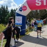 Mężczyzna w niebieskiej koszulce przekracza linię mety. Tło stanowią zielone drzewa i niebo z białymi chmurami. Grupa ludzi klaska na poboczu.