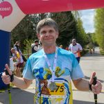 Uśmiechnięty mężczyzna z kijkami stoi przed różowym balonem startowym. Nosi kolorową koszulkę z numerem 175. W tle znajdują się drzewa i inni uczestnicy wydarzenia.