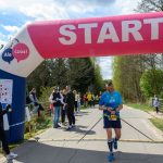 Zdjęcie przedstawia biegacza z kijkami, startującego pod różowo-białym balonem z napisem „START”. W tle jest grupa kibiców na drodze otoczonej drzewami. Na niebie są białe chmury, a słońce świeci jasno.