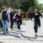 Dwóch biegaczy w czarnych strojach przekracza linię mety na zewnętrznej drodze. W tle zielone drzewa i widzowie. Jeden z widzów trzyma kartki i ma na sobie czapkę i kamizelkę.