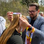 Na zdjęciu mężczyzna zakłada medal na szyję młodej kobiety. Tło stanowi las, a atmosfera jest słoneczna. Medale mają jasne kolory z napisami.