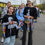 Grupa młodych osób trzyma medale, stojąc na asfaltowej drodze. Tło to zielone drzewa i niebo z chmurami. Dominują ciemne i jasne ubrania.