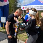 Biegacz otrzymuje medal od kobiety przy mecie. Tło zielone z niebieskim namiotem i zegarem z czasem 0:42:16.