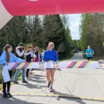 Zawodniczka w niebieskiej kurtce przecina linię mety. Nad nią kolorowy łuk, tło to drzewa i niebo. Wokół stoją kibice.