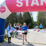 Biegaczka przekracza linię mety pod różowo-białym łukiem startowym. Na poboczu stoją ludzie i kibicują. W tle drzewa i asfaltowa droga.