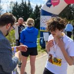 Grupa ludzi po biegu, w tle brama z napisem. Mężczyzna wręcza medale, a uczestnik w białej koszulce poprawia naszyjnik. Pogoda słoneczna, drzewa w tle.