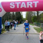 Biegaczka w niebieskim stroju przekracza linię startu pod różowo-białym łukiem. W tle drzewa i osoby obserwujące. Elektroniczny zegar pokazuje czas 0:43:25.