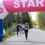 Biegaczka w czarnym stroju przekracza linię startu pod różowym banerem „START”. W tle drzewa i kibice. Pogoda jest słoneczna.