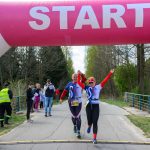 Dwóch biegaczy w sportowych strojach przekracza linię startu pod różowo-białym balonem. W tle drzewa i kibice. Atmosfera radości i energii.