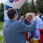 Mężczyzna zakłada medal biegaczowi na mecie. W tle widać las i kilku obserwatorów. Ubrania w głównie niebiesko-białych kolorach.