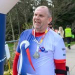 Mężczyzna w sportowym stroju z medalem stoi na zawodach biegowych. Ubrany w biało-niebieską koszulkę z czerwonymi akcentami. W tle widoczne są drzewa i ludzi.