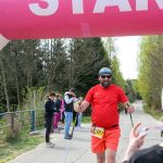 Zawodnik w czerwonym stroju kończy marsz nordic walking pod różowym balonem START na leśnej drodze. W tle inni uczestnicy i drzewa. Pogoda jest słoneczna.