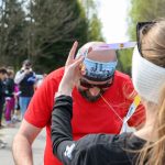 Na zdjęciu widać kobietę zakładającą medal mężczyźnie w czerwonej koszulce podczas biegu. W tle rozmyte drzewa i uczestnicy. Mężczyzna ma opaskę, kobieta nosi białą przepaskę na włosach.