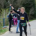 Na zdjęciu kobieta z numerem startowym 186 kończy nordic walking. Tło to drzewa i grupa ludzi. Przewaga kolorów zielonego i czarnego.