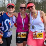 Trzy uśmiechnięte osoby w sportowych strojach ze srebrnymi medalami. Kolory: różowy, biały, niebieski. W tle drzewa i uczestnicy biegu.