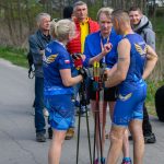 Na zdjęciu grupa osób w sportowych strojach w niebieskim kolorze stoi na drodze. Trzymają kijki do nordic walking, a w tle widać drzewa. Atmosfera jest luźna, a w tle są inni uczestnicy.