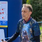 Na zdjęciu mężczyzna w koszulce z motywem wilka. W tle znajduje się plakat na niebieskim tle. Otoczenie jest zielone, z drzewami i płotem.