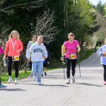 Grupa ludzi biegnie po brukowanej ścieżce przez las. Kobieta w różowej koszulce używa kijków do nordic walking. Tło to zieleń drzew i jasne niebo.