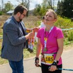 Kobieta w różowej koszulce z medalem stoi na trasie biegu. Mężczyzna w marynarce wręcza medale. W tle wiosenna sceneria i mapa.