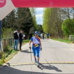 Zawodniczka na trasie biegu w niebieskiej odzieży sportowej. W tle wiosenna zieleń drzew. Napis Ale czas! na łuku nad drogą.