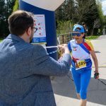 Starsza kobieta w niebieskim stroju biegnie na zawodach. Mężczyzna w marynarce wręcza jej kartkę. W tle las i droga.