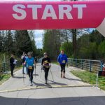 Trzy osoby z kijkami przechodzą pod różową bramą startową. W tle drzewa i błękitne niebo. Obok bramy licznik czasu.