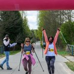 Dwie kobiety z kijkami przekraczają linię startu na zawodach nordic walking. Tło to zielony las i droga. Nad nimi różowy napis START.